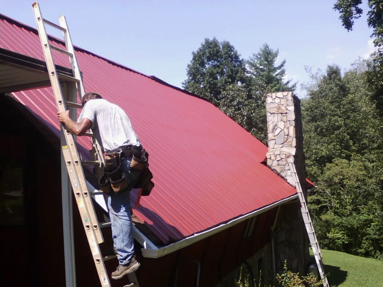 Roofer on a ladder beside a red metal roof and stone chimney — Soffit & Fascia Repair by Roofing Brothers in Hunlock Creek